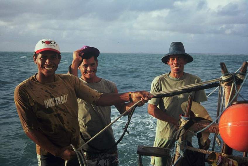 Three jangadeiros on a jangada. One of them is steering while the other two hold espeque lines.