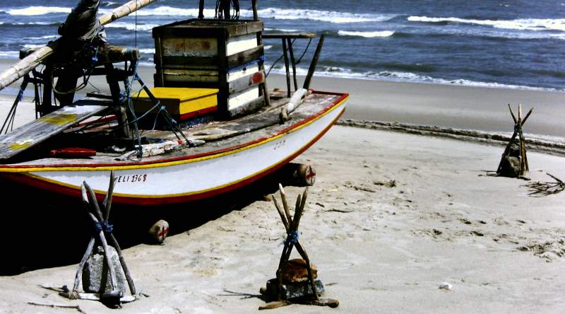A jangada on the beach with three stone anchors sitting on the sand beside it.