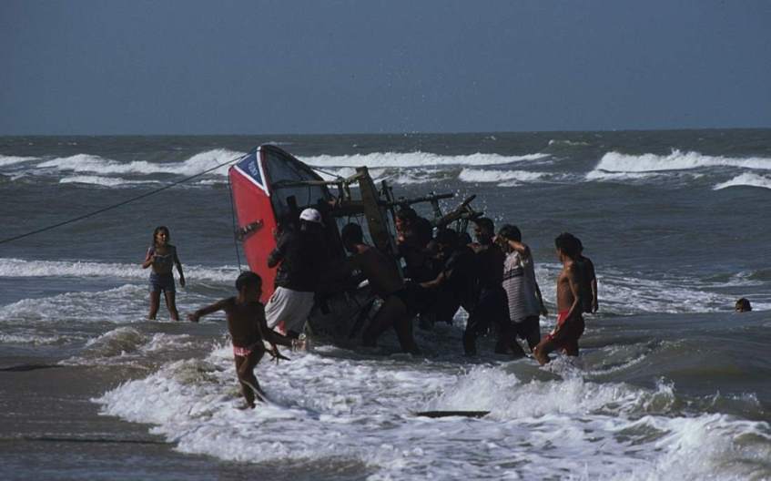 A jangada on it's side in the surf, being righted by a group of people.