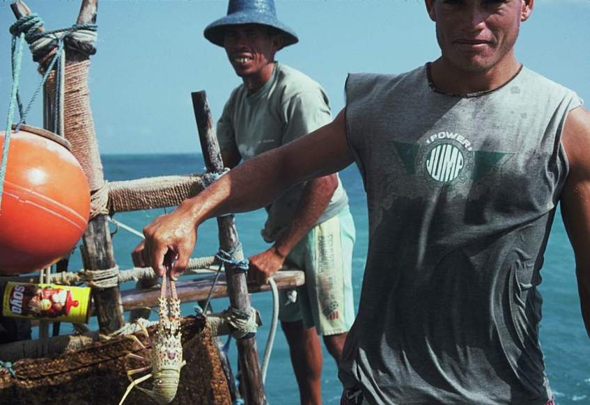 Jangadeiro steering while another holds up a lobster he just caught.